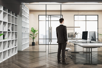 Fototapeta premium Attractive thoughtful european businessman standing in wooden and concrete office interior with furnituire and window with city view. CEO, executive and consulting concept.