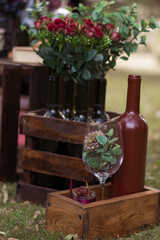 A brown bottle of wine next to a transparent glass goblet stands in a wooden box. Flowers inside the glass. Roses in bottles in the background