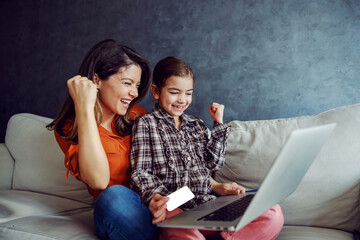 Smiling mother and daughter sitting on sofa and using laptop for online shopping. Daughter holding credit card. They had successful purchase so they are overjoyed and holding fists.
