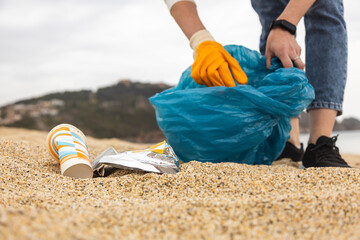 A woman in gloves with a special blue bag picks up garbage among the sand along the coast. The problem of environmental pollution. Cleaning up trash on the beach