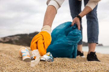 A woman in gloves with a special blue bag picks up garbage among the sand along the coast. The...