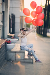 Funny stylish European girl child with red balloons in torn jeans sits on stairs on sunny summer day. Child in sunglasses at sunset on city streets generation Z, happy birthday