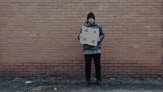 Homeless holds piece of cardboard with markers for planar tracking. Refugee is at brick wall looking for job. Male tramp in dirty clothes. Below poverty line. Bearded fugitive beggar. Drunk immigrant