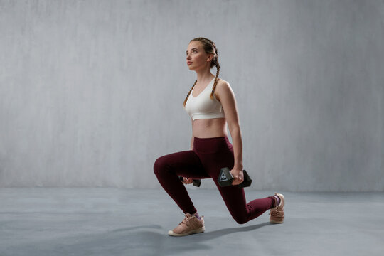 Young athletic woman in sportswear doing squat using dumbbells, posing in studio with grey background.