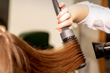 A hairdresser is drying long brown hair with a hairdryer and round brush in a beauty salon