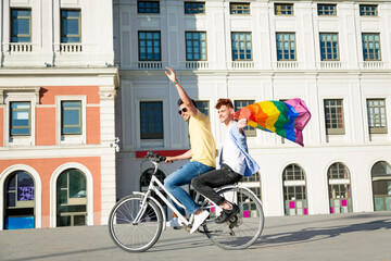 Fototapeta na wymiar Young gay men couple having fun riding bicycles with gay pride flag outdoors. lgbt concept