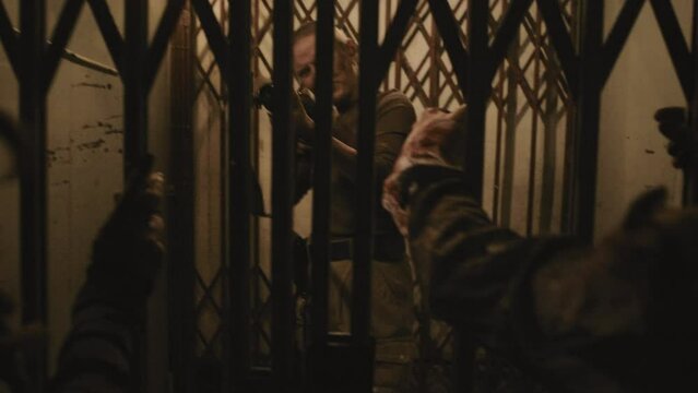 Medium long of young Caucasian woman with shaved head shooting zombies behind old elevator gate in creepy old building