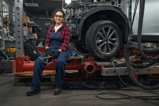 Cheerful auto mechanic sitting by car in vehicle repair shop