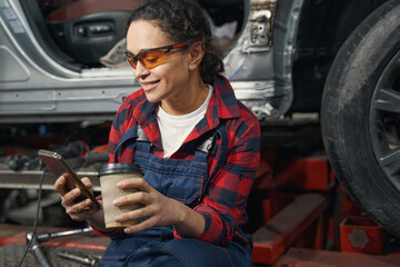 Woman auto mechanic using cellphone and drinking coffee at work