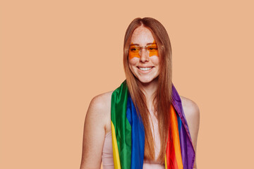 Lgbtq concept. Positive caucasian girl wears stylish sunglasses, holding rainbow flag isolated in studio over beige background.