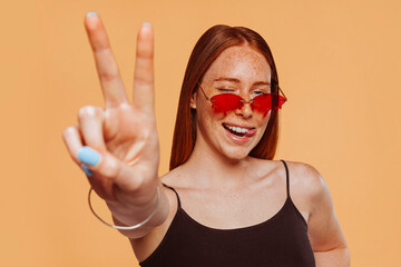 Joyful caucasian woman with peace hand sign, blinks eye and shows tongue, has straight hair, dressed casually, wears red stylish sunglasses, poses against beige background.