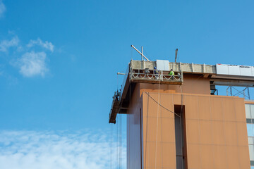 Using a construction gondola lift, workers place panels on a towering skyscraper. Building construction with blue sky as background