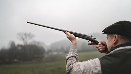 Hunter man in traditional shooting clothes on field aiming with shotgun.