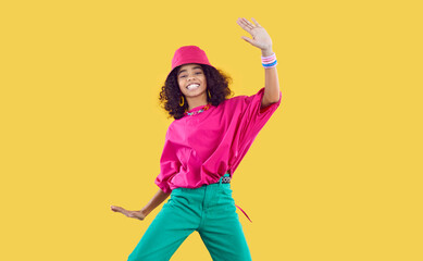 Happy joyful Black kid in funky outfit dancing in studio. Cheerful African dancer girl wearing loose fuchsia top, bucket hat and green pants enjoying hype and having fun. Children's fashion concept
