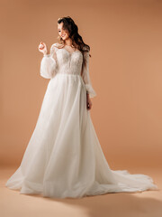 Fototapeta na wymiar Young brunette bride in elegant white lace wedding dress with long sleeves and lush tulle skirt, big train. Full-length portrait of happy smiling bride in studio. Bridal fashion. Wedding inspiration