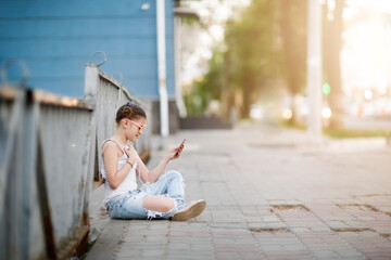 pretty girl with headphones and a phone sits on the sidewalk, a lonely child on a city street....