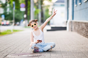 pretty girl with headphones and a phone sits on the sidewalk, a lonely child on a city street....