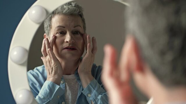 An elderly graying woman carefully examines her reflection in the mirror, lightly touching the wrinkles on the skin of her face.