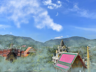 Thick fog stretches in the valley between the green hills. Buddhist monastery, immersed in clouds of fog. Bright roofs of religious buildings and spiers. Blue sky with clouds.  Crescent moon. Thailand