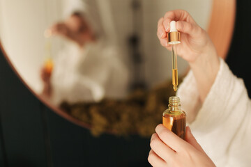 A woman in a white bathrobe and with a bath towel is using a pipette to apply an oily serum for skin