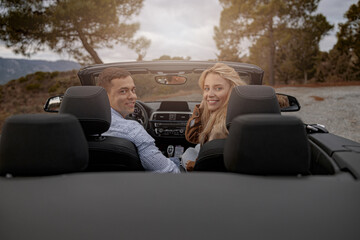 Happy couple of man and woman sitting in convertible car smiling while looking back, vacation trip - obrazy, fototapety, plakaty