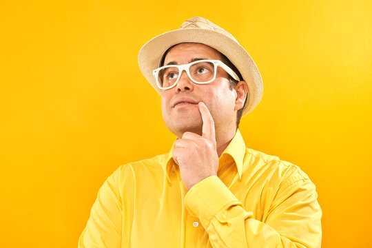 Positive man in panama hat holding chin thinks about vacation, doubts, makes decision isolated on yellow studio background