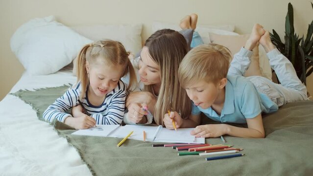 Family leisure, mom and kids lay on bed and draw pictures on paper in their apartment room. Educational games, happy friendly family, parents and children relationships