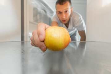 Man takes out a single apple from an empty refrigerator. Concept of delivery service, hunger, dients . Photo from inside the refrigerator - obrazy, fototapety, plakaty