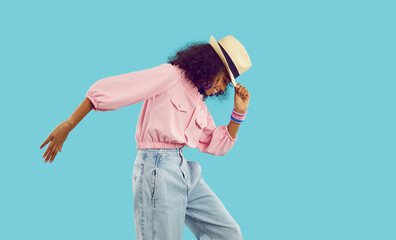 Happy child having fun in fashion studio. Funny little kid in casual summer fit dancing isolated on blue background. Profile side view African American girl in pink top, hat and jeans doing moonwalk