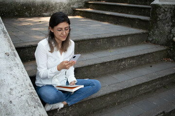 Fototapeta na wymiar YOUNG MILLENNIAL GIRL TEXTING ON HER CELL PHONE WHILE READING A BOOK IN THE PARK