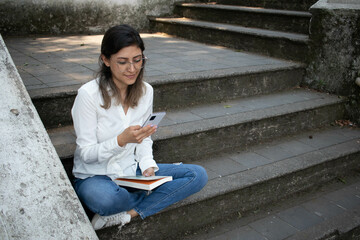 YOUNG MILLENNIAL GIRL TEXTING ON HER CELL PHONE WHILE READING A BOOK IN THE PARK