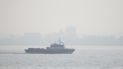 Fototapeta premium Boat sailing on a misty morning close to the shore against an urban city background.