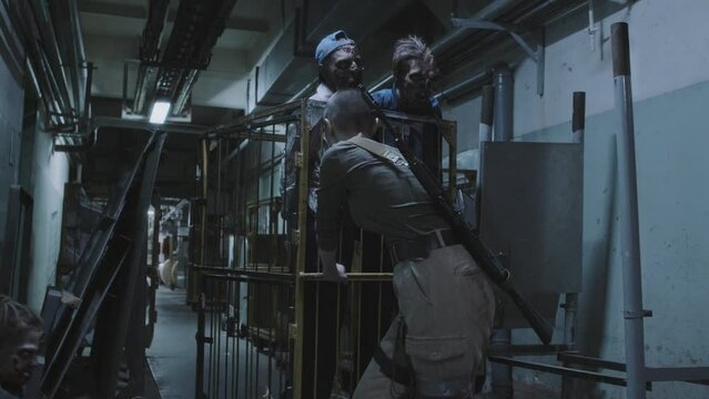 Injured female soldier with shaved head transporting zombies in cage on wheels in dark basement of industrial building