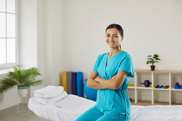 Portrait of friendly confident professional female masseur or chiropractor in her medical office. Young woman in blue medical uniform sits on massage couch with folded arms and looks at camera.