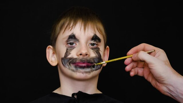 portrait of a caucasian boy 8-9 years old who is applied by a brush with a creepy make-up of a zombie monster preparing for a masquerade. a child in black clothes in the form of a horror story