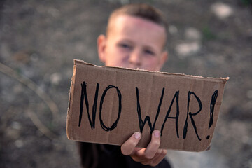 A boy with a grimy face and sad eyes holds a cardboard poster with the inscription NO WAR