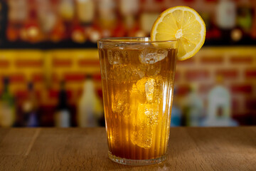 Glass with sparkling cola, ice and lemon on a wooden counter in front of a bar