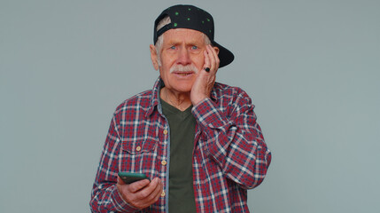 Senior man in shirt use mobile cell phone typing browsing, loses becoming surprised sudden lottery results, bad fortune, loss, fail. Elderly grandfather isolated alone on gray studio wall background