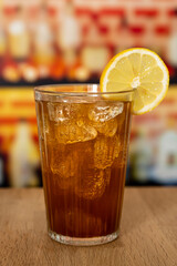 Glass with sparkling cola, ice and lemon on a wooden counter in front of a bar.