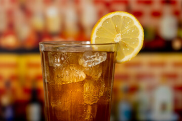 Glass with sparkling cola, ice and lemon on a wooden counter in front of a bar