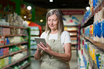 Portrait of senior female consultant, employee in grocery store, supermarket. Stands with a tablet,...