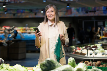 Senior beautiful woman shopping for vegetables in the store. Supermarket. He stands, holds the phone in his hands, holds the food net, chooses. He looks at the camera, smiles