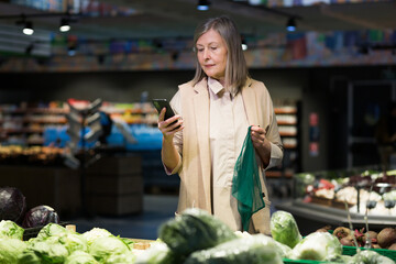 Senior beautiful woman shopping for vegetables in the store. Supermarket. He stands, holds the phone in his hands, holds the food net, chooses. He looks at the camera, smiles