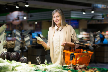 Portrait. Senior beautiful woman shopping for vegetables in the store. Supermarket. Stands, holds a list of products written on paper in his hands,  chooses. looks at the camera, smiles