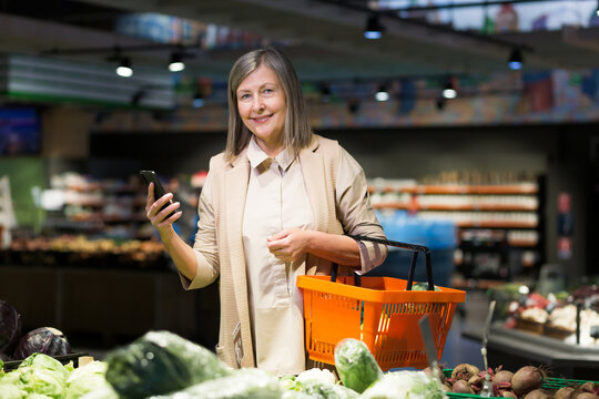 Portrait. Senior beautiful woman shopping for vegetables in the store. Supermarket. He stands, holds the phone in his hands, holds a plastic food basket, chooses. She looks at the camera, smiles - Powered by Adobe