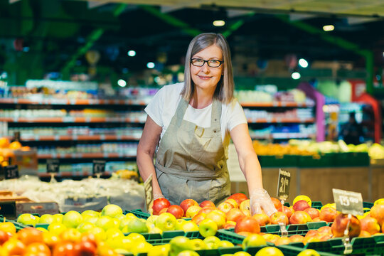 Portrait of senior woman in glasses at grocery store employee, supermarket. Stands in work clothes, near fruits and ofochi, shows food, looks at the camera, smiles