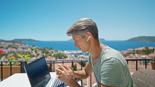 dolly zoom out footage man in earphones working with laptop outdoors by beautiful blue sea view, speaking by online video call. Concept remote work and travel.