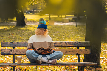 Attractive blond curly haired girl reads a book on the beach in park, drinking coffee autumn copy space full shot . High quality photo