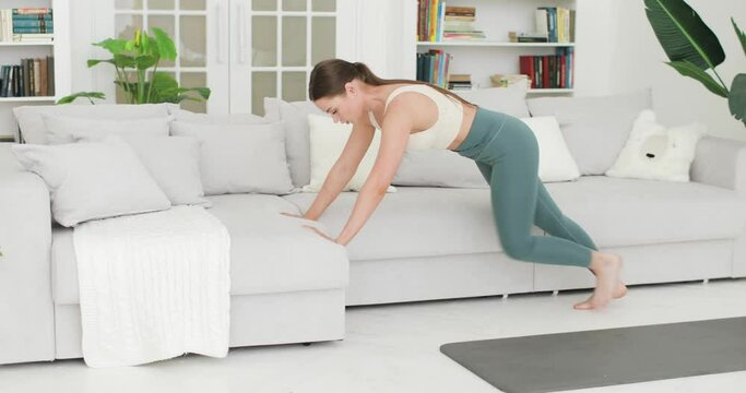 Young brunette woman is having workout. Woman is training bending the legs alternately at home in living room. She does fitness exercise. Workout, sport, fitness, training and wellness concept.