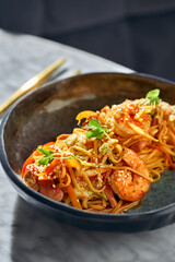 Stir fry noodles with vegetables and shrimps and seafood in black bowl on a marble table.